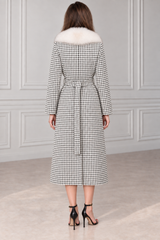 A woman viewed from the back wearing a long black and white houndstooth coat with a white fur collar and a matching waist tie, paired with black high-heeled sandals.
