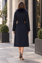 A woman viewed from behind wearing a dark navy wool coat with a dark fur collar, a tan belt, and black high heels, standing on a city sidewalk.