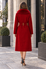 A woman viewed from behind wearing a bright red knee-length coat with a fur-like collar and a matching belt with gold-tone hardware, standing on a city sidewalk in front of a stone building.