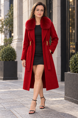 A woman wearing a long red coat with a large dark fur collar, a black dress, and black heeled sandals, standing outdoors on a city sidewalk.