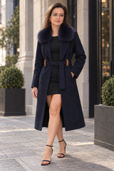 A woman stands outdoors wearing a long navy wool-blend coat with a large dark fur collar, a matching waist tie belt, and brown leather accents, over a black dress and black strappy heels.