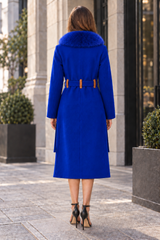 A woman viewed from behind wearing a bright royal blue mid-length coat with a fur-trimmed collar and a matching belt with gold-tone hardware, standing on a city sidewalk.