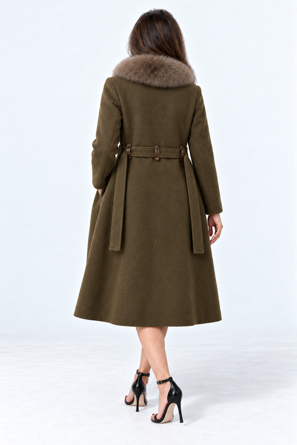 A woman viewed from the back wearing an olive green belted coat with a brown fur collar, paired with black high-heeled sandals, against a light gray studio background.