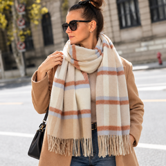 A woman wearing a large cream-colored plaid scarf with brown and light blue stripes, styled with a tan coat, sunglasses, and a black handbag in an outdoor urban setting.
