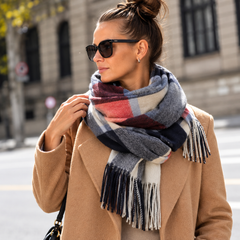A woman wearing a thick plaid scarf in shades of gray, navy, red, and cream, styled with a camel coat and sunglasses in an outdoor urban setting.