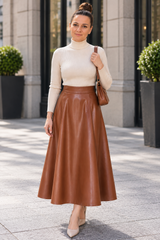 A woman stands outdoors on a city sidewalk wearing a cream ribbed turtleneck sweater and a brown faux-leather midi skirt, carrying a brown shoulder bag.