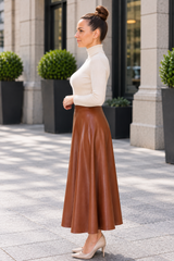 A woman stands outdoors in a city setting wearing a cream mock-neck long-sleeve top and a brown faux-leather midi skirt, paired with beige pointed-toe heels.