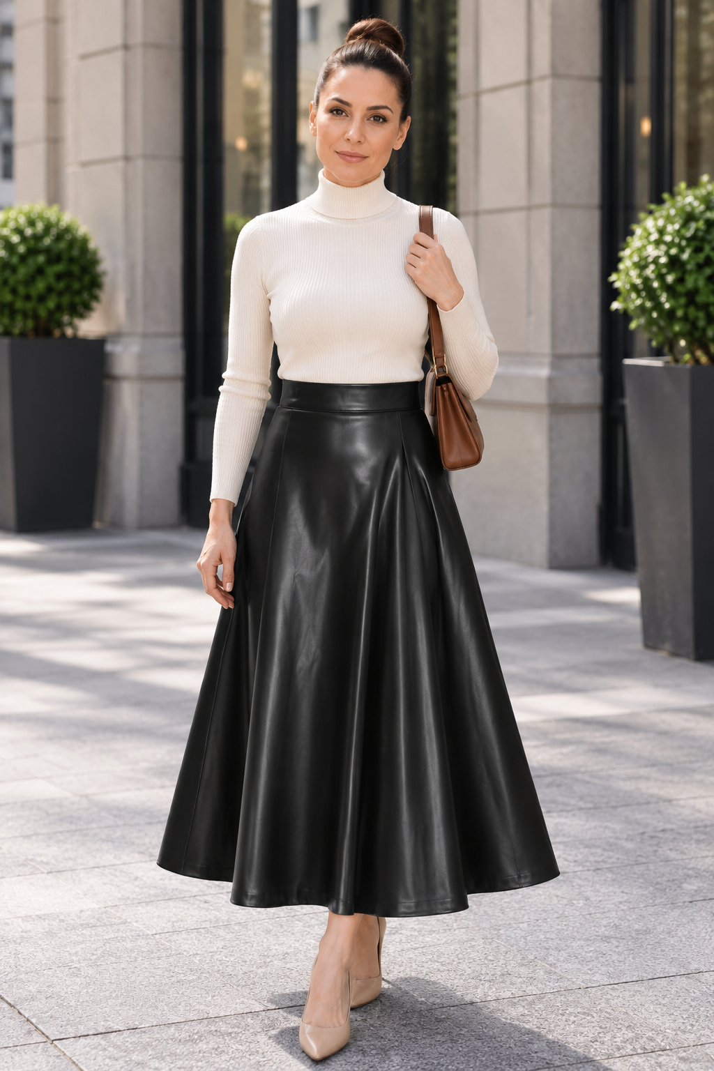 A woman stands outdoors on a city sidewalk wearing a cream mock-neck long-sleeve top and a black faux-leather midi skirt, carrying a brown shoulder bag.