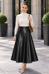 A woman stands outdoors on a city sidewalk wearing a cream mock-neck long-sleeve top and a black faux-leather midi skirt, carrying a brown shoulder bag.