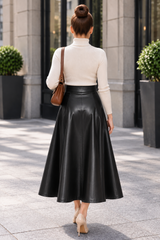 A woman viewed from behind wearing a cream mock-neck long-sleeve top and a high-waisted black faux-leather midi skirt, carrying a brown shoulder bag while walking on a stone sidewalk.