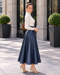 A woman stands outdoors in front of a modern building, wearing a fitted white mock-neck long-sleeve top and a high-waisted navy blue midi skirt. She is carrying a brown shoulder bag and wearing beige high-heeled pumps.
