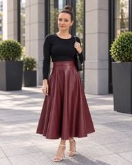 A woman stands outdoors in front of a modern building, wearing a black long-sleeve top and a high-waisted, flared burgundy faux-leather midi skirt. She is carrying a black shoulder bag and wearing clear high-heeled sandals.