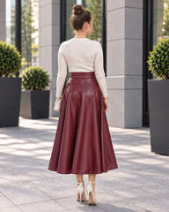 A woman viewed from behind wearing a cream long-sleeve knit top and a high-waisted, flared burgundy faux-leather midi skirt, standing on a paved outdoor walkway.