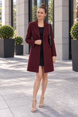 A woman stands outdoors in front of a modern building, wearing a long burgundy blazer over a black dress, with white heeled sandals and a black shoulder bag.