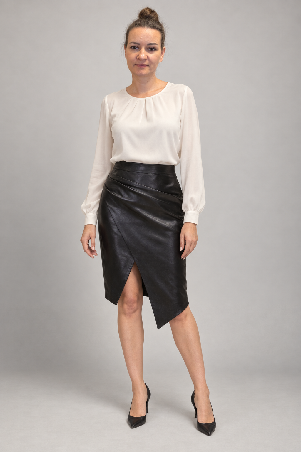 A woman modeling a black faux-leather wrap skirt with a side slit and an asymmetrical hem, paired with a cream long-sleeve blouse and black heels, against a solid gray studio background.