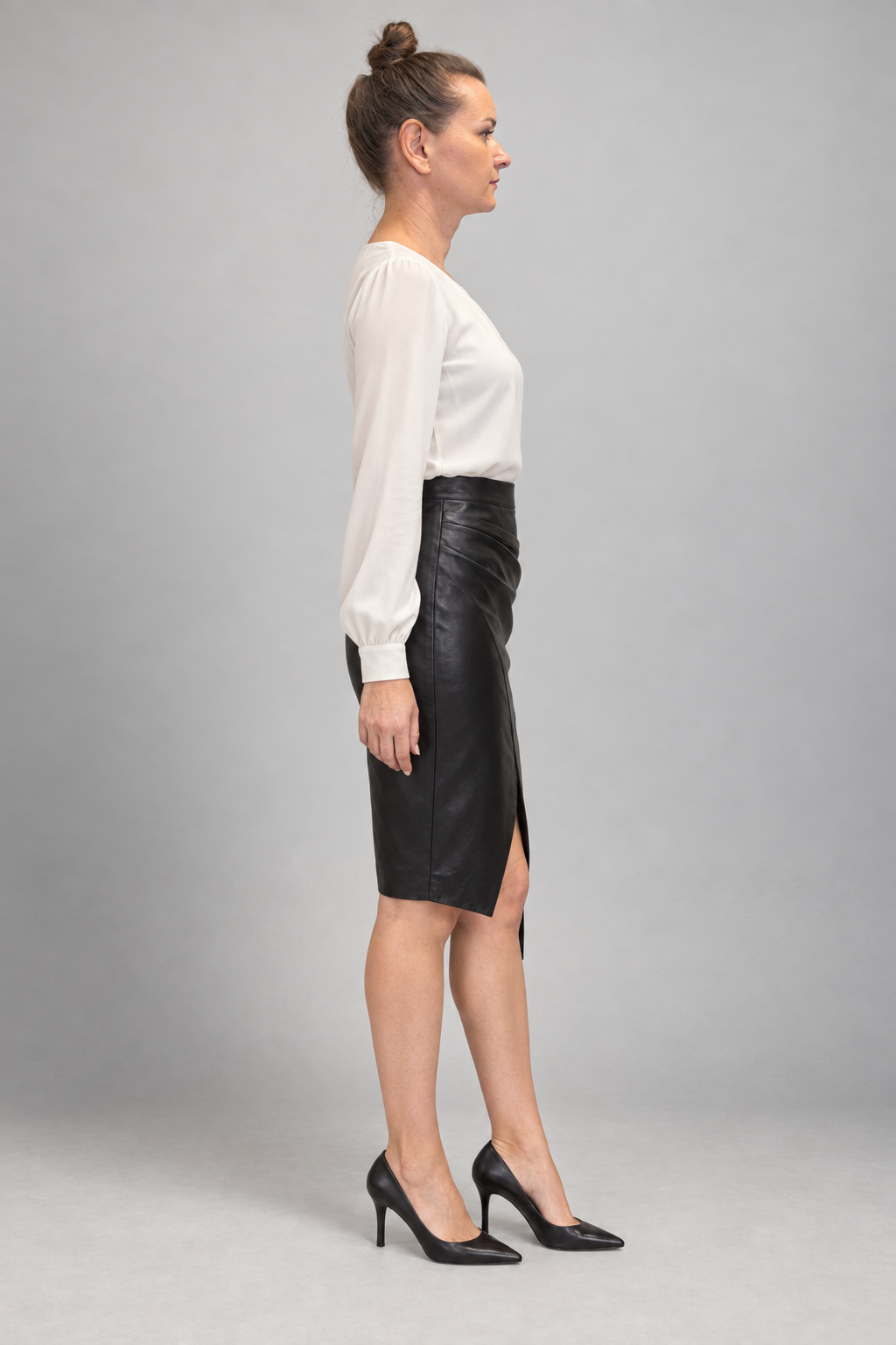A woman in side profile wearing a white long-sleeve blouse, a black faux-leather pencil skirt with a front slit, and black pointed-toe heels, standing against a neutral gray studio background.
