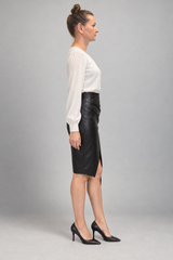 A woman in side profile wearing a white long-sleeve blouse, a black faux-leather pencil skirt with a front slit, and black pointed-toe heels, standing against a neutral gray studio background.