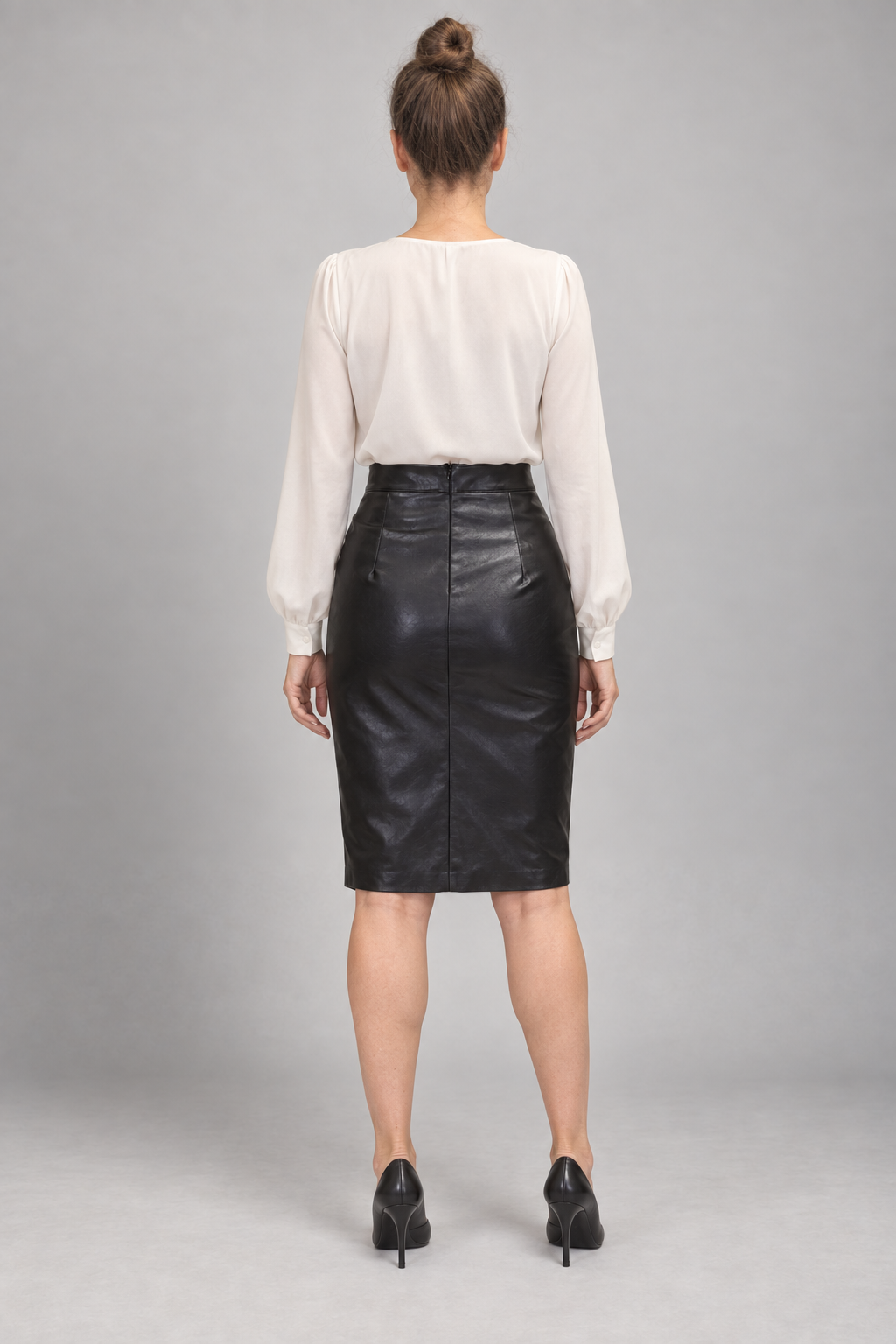 A woman shown from the back wearing a cream-colored long-sleeve blouse and a black faux-leather pencil skirt, paired with black heels, against a neutral gray studio background.