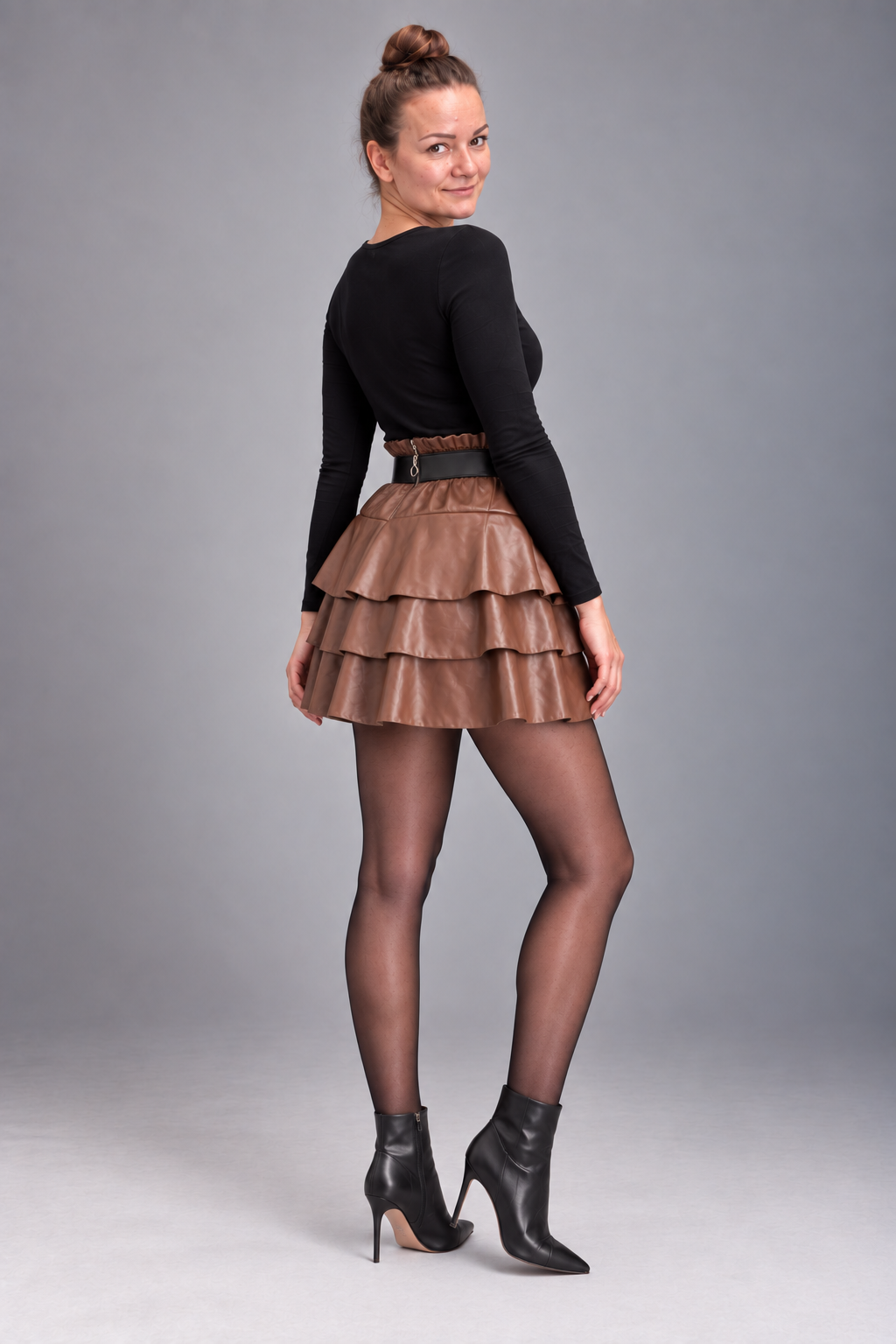 A woman modeling a black long-sleeve top, a brown tiered faux-leather mini skirt with a matching belt, sheer black tights, and black ankle boots, photographed against a solid gray studio background.