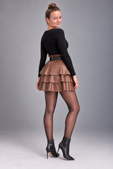 A woman modeling a black long-sleeve top, a brown tiered faux-leather mini skirt with a matching belt, sheer black tights, and black ankle boots, photographed against a solid gray studio background.