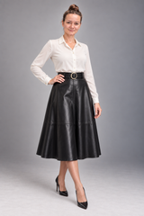 A woman modeling a white long-sleeve button-up shirt and a black high-waisted faux-leather A-line midi skirt with a black belt featuring a gold-tone round buckle, paired with black pointed-toe heels, against a solid gray studio background.