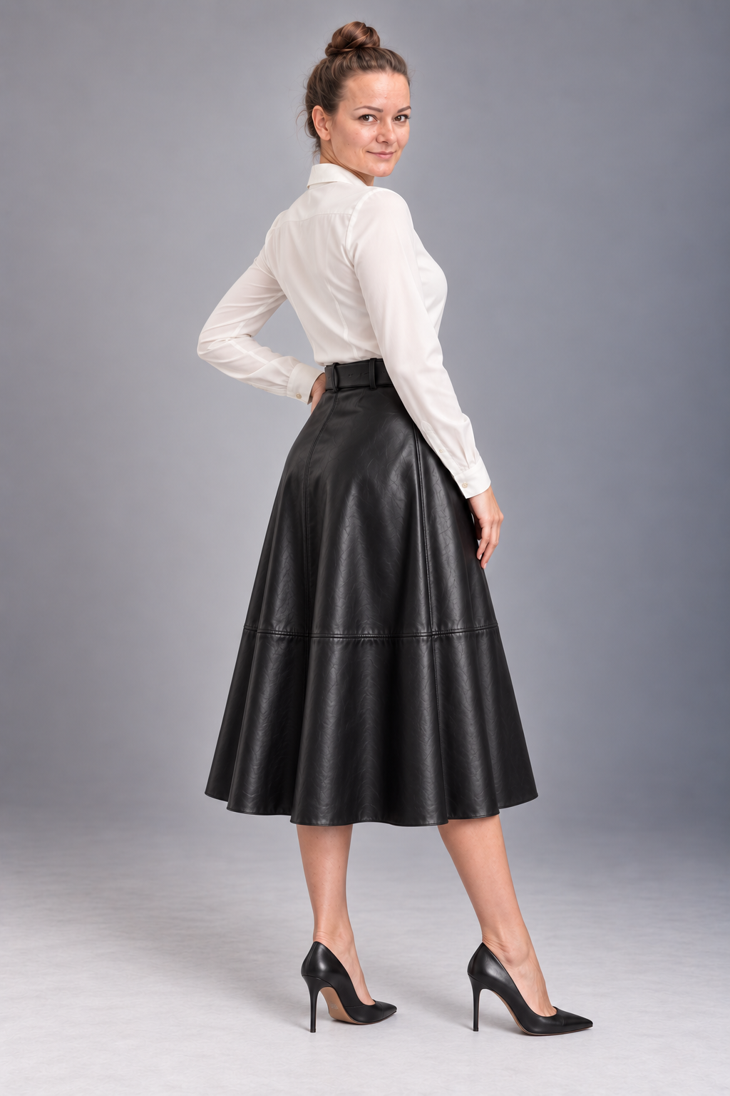 A woman modeling a black faux-leather midi skirt with a high waist and flared A-line silhouette, paired with a white long-sleeve blouse and black pointed-toe heels, photographed against a neutral gray studio background.