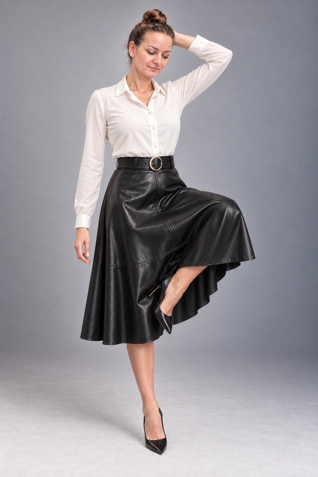 A woman modeling a high-waisted black faux-leather midi skirt with a matching black belt and gold buckle, paired with a white button-down shirt and black heels, against a gray studio background.