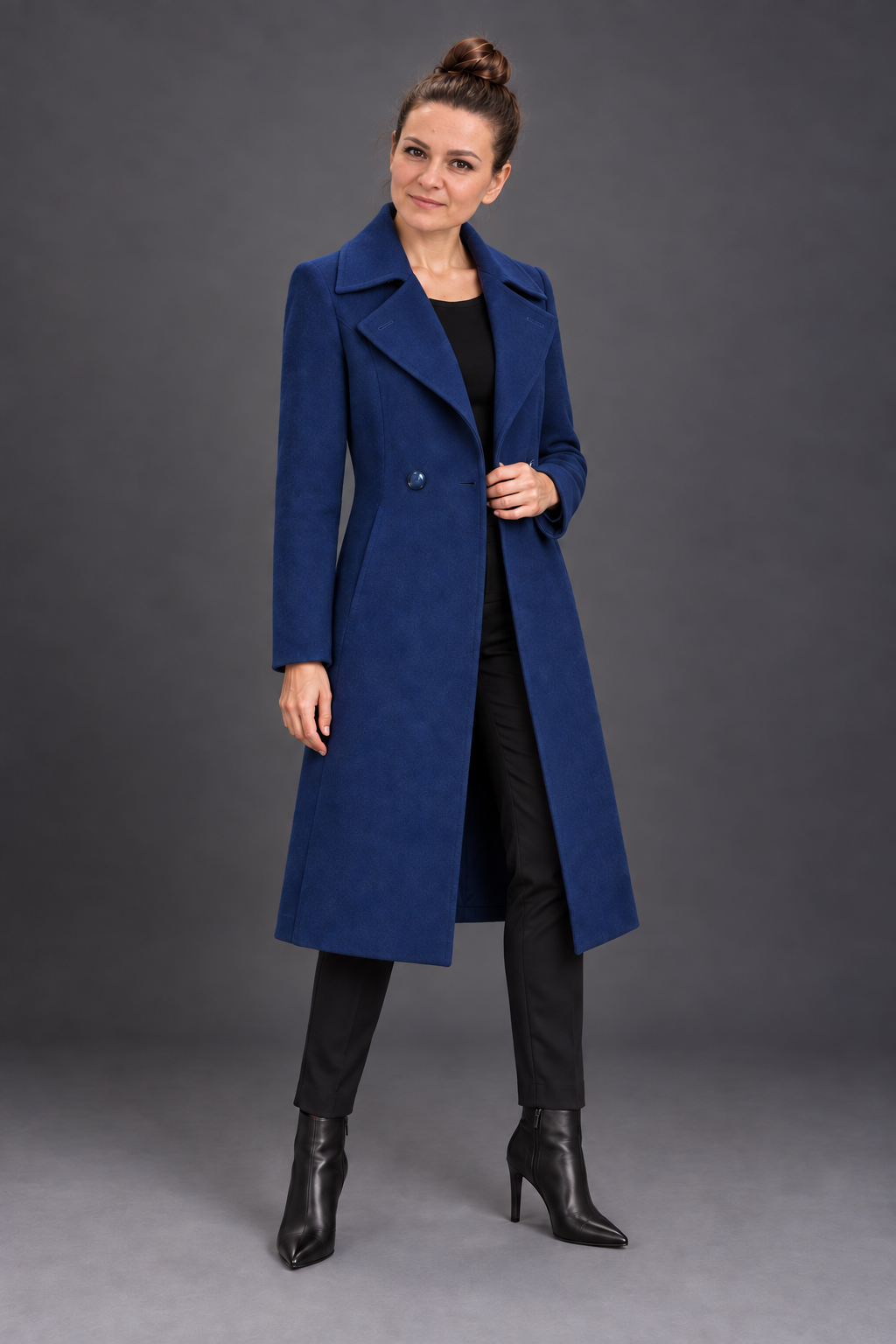 A woman modeling a long, double-breasted blue coat over a black outfit, standing against a dark gray studio background.