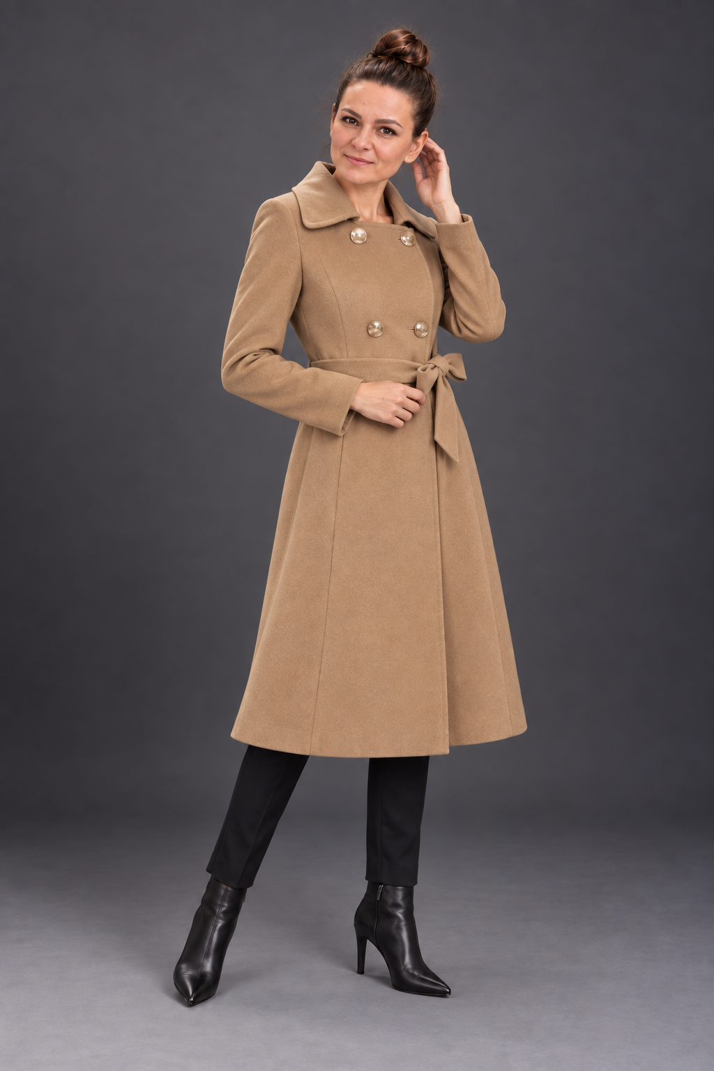 A woman modeling a mid-length camel brown double-breasted coat with a tie waist belt, paired with black pants and black ankle boots, against a dark gray studio background.