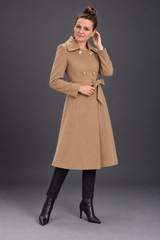 A woman modeling a mid-length camel brown double-breasted coat with a tie waist belt, paired with black pants and black ankle boots, against a dark gray studio background.