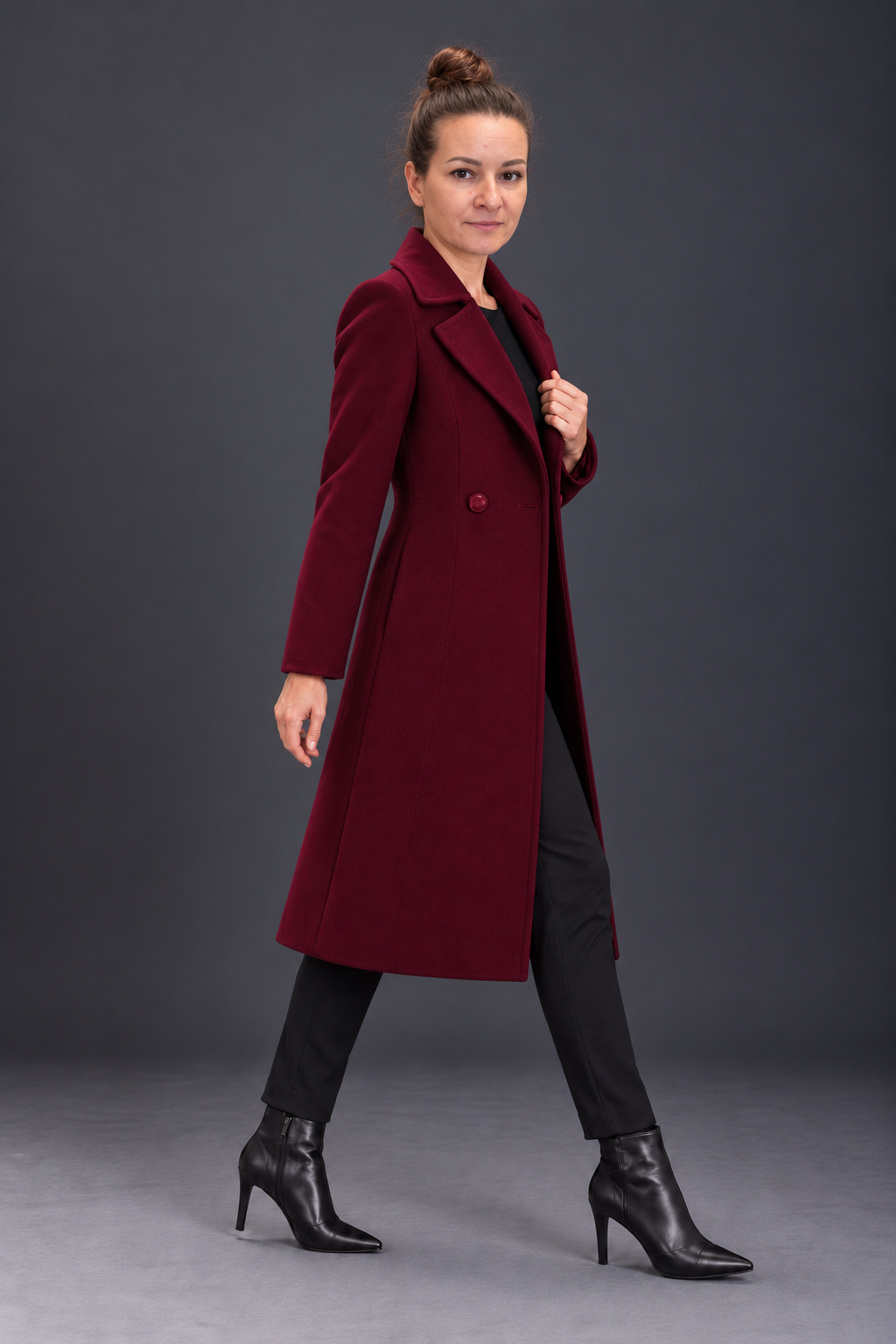 A woman modeling a long burgundy wool-blend coat over a black outfit, standing against a dark gray studio background.