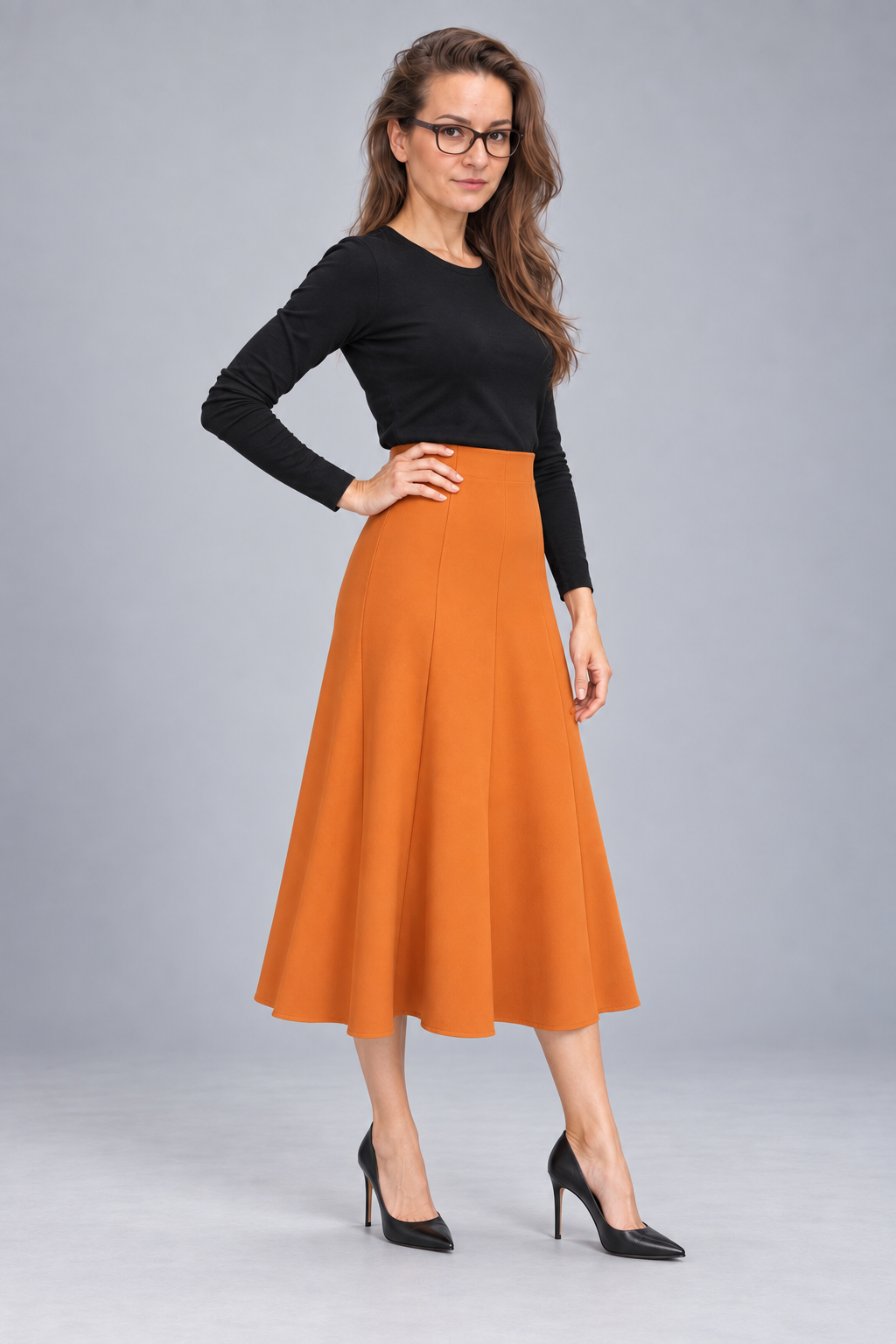 A woman modeling a black long-sleeve top and a high-waisted, pleated orange A-line midi skirt, paired with black pointed-toe heels, against a solid gray studio background.