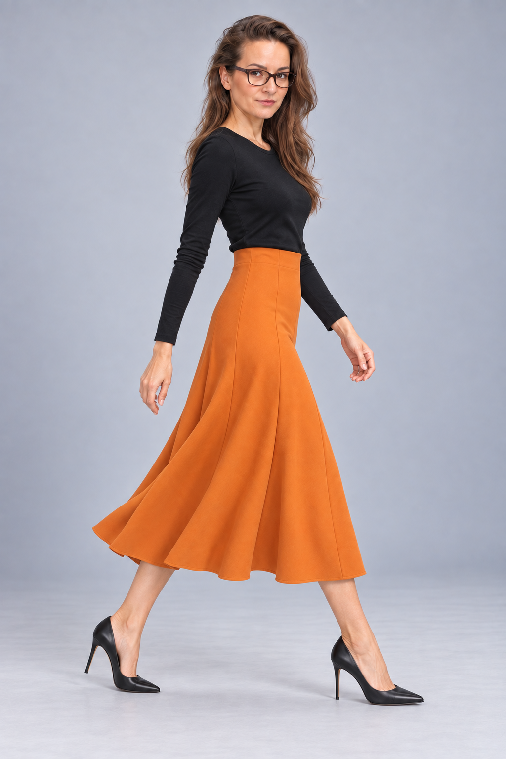 A woman wearing a black long-sleeve top, a high-waisted orange A-line midi skirt, and black pointed-toe heels, posing against a solid light gray studio background.