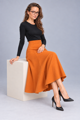 A woman wearing a black long-sleeve top and a high-waisted orange flared midi skirt, sitting on a white cube against a light gray studio background.