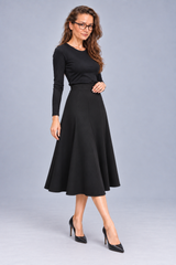 A woman modeling a black long-sleeve midi dress with a flared skirt, paired with black pointed-toe heels, against a solid light gray studio background.