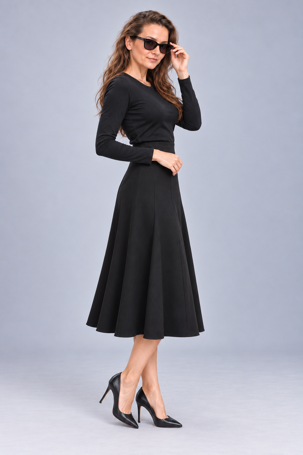 A woman modeling a black long-sleeve midi dress with a flared skirt, paired with black pointed-toe heels and sunglasses, against a solid light gray studio background.
