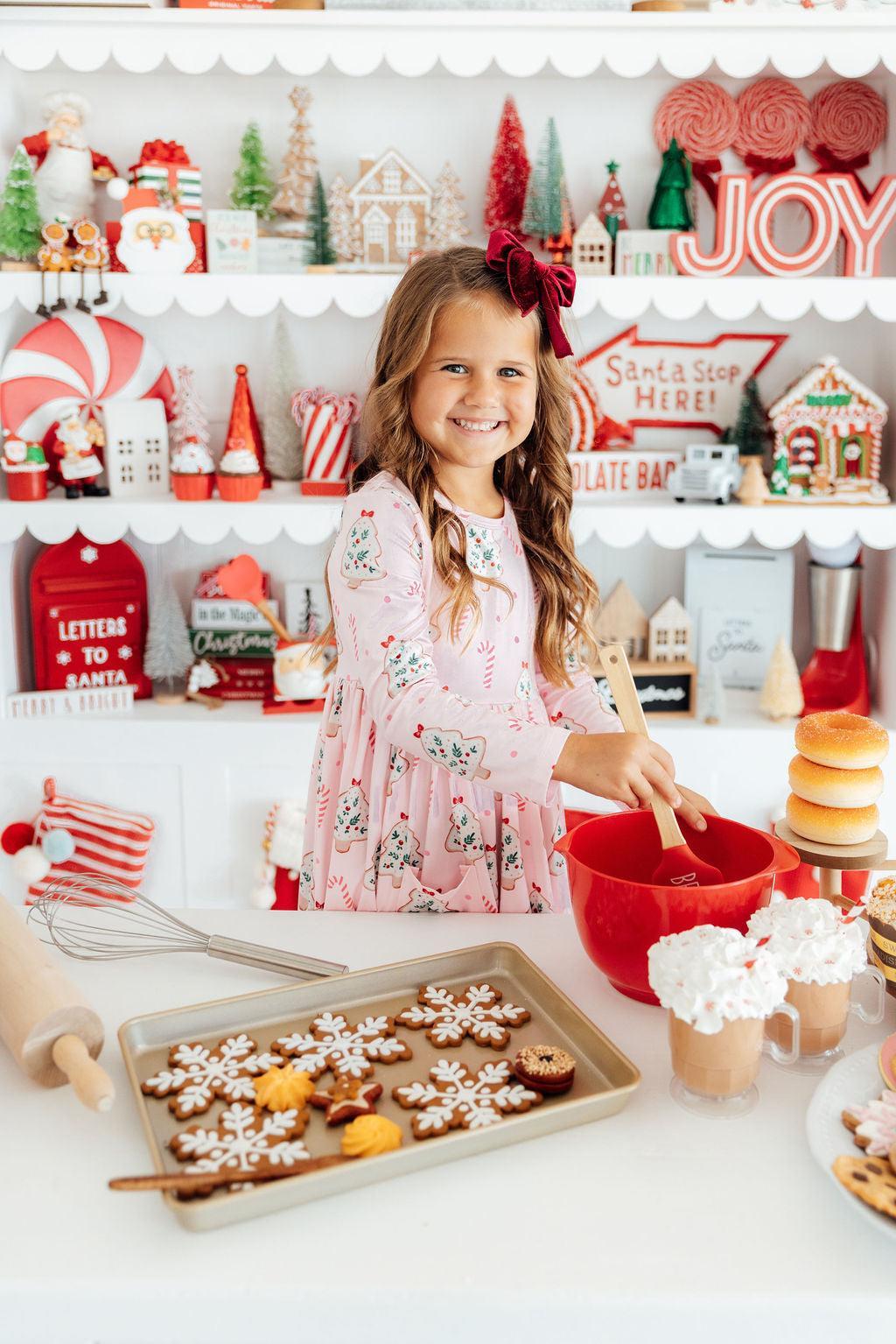 A young girl with a red bow in her hair smiles while mixing ingredients in a red bowl at a festive holiday kitchen table. A baking tray of decorated gingerbread cookies sits in the foreground, with shelves of Christmas decorations and signs in the background.