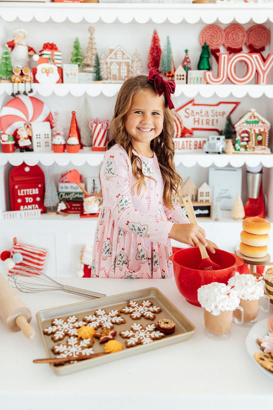 A young girl with a red bow in her hair smiles while mixing ingredients in a red bowl at a festive holiday kitchen table. A baking tray of decorated gingerbread cookies sits in the foreground, with shelves of Christmas decorations and signs in the background.