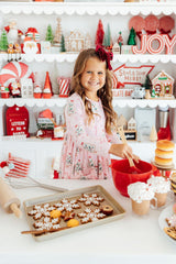 A young girl with a red bow in her hair smiles while mixing ingredients in a red bowl at a festive holiday kitchen table. A baking tray of decorated gingerbread cookies sits in the foreground, with shelves of Christmas decorations and signs in the background.