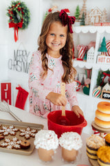 A young girl with a red bow in her hair smiles while mixing ingredients in a red bowl at a decorated Christmas table. She is wearing a pink holiday print pajama shirt.