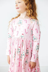 A young girl with curly light brown hair wearing a pink long-sleeve dress with a festive gingerbread cookie and candy cane print, photographed against a clean white background.