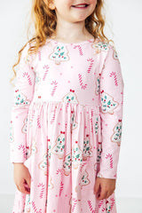 A young girl wearing a pink long-sleeve dress with a festive Christmas print of gingerbread cookies, candy canes, and snow globes, shown against a plain white background.