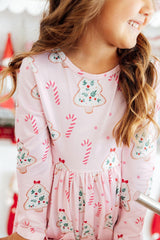 A close-up of a young girl wearing a light pink long-sleeve dress with a festive Christmas print featuring gingerbread trees, candy canes, and snowman motifs.