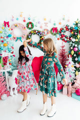 Two children in festive dresses standing in front of a colorful Christmas-themed backdrop with wreaths and decorations.