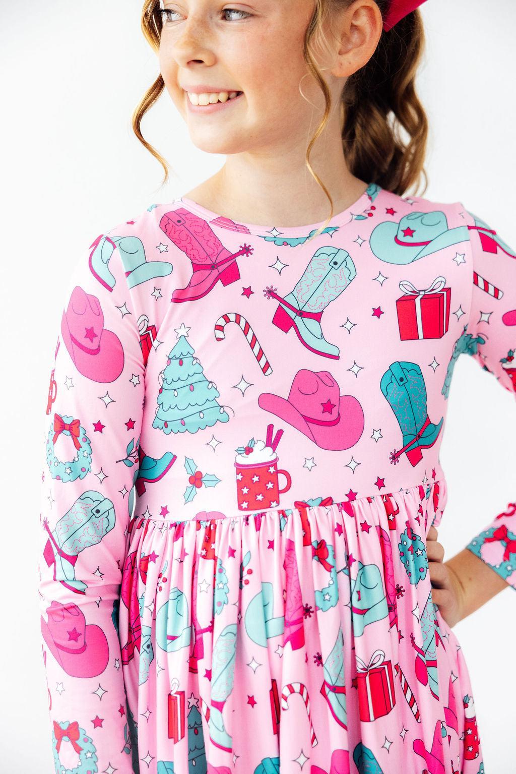 A young girl wearing a pink long-sleeve dress with a cowboy and holiday print, featuring pink and teal cowboy hats, boots, a Christmas tree, candy canes, and hot cocoa mugs, set against a plain white background.