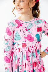 A young girl wearing a pink long-sleeve dress with a cowboy and holiday print, featuring pink and teal cowboy hats, boots, a Christmas tree, candy canes, and hot cocoa mugs, set against a plain white background.