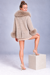 A woman models a beige faux-fur jacket with a large matching collar and cuffs, paired with a short beige dress and nude high heels, standing on a light gray pedestal against a gray studio background.