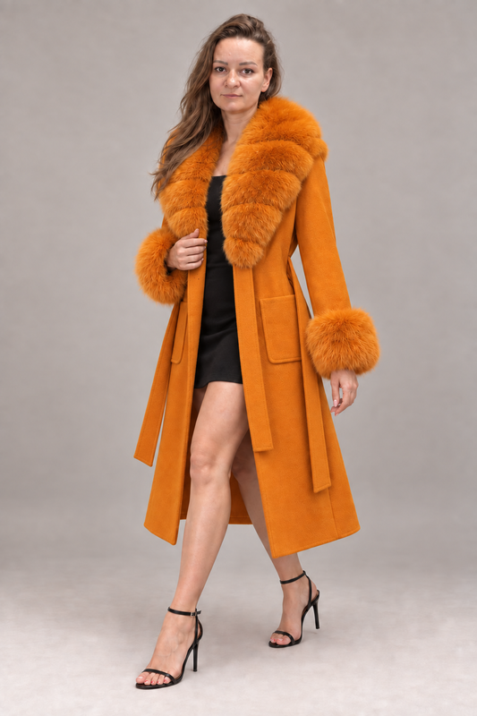 A woman modeling a long orange coat with a large orange fur collar, cuffs, and hem, worn over a black dress and black high-heeled sandals, against a neutral gray studio background.