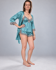 A woman modeling a satin sleepwear set consisting of a teal blue robe with silver lace trim, a matching lace-trimmed camisole, and matching lace-trimmed shorts, against a light gray studio background.