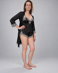 A woman modeling a black satin lingerie set with silver lace trim, consisting of a long-sleeve robe and matching thong, against a neutral gray studio background.